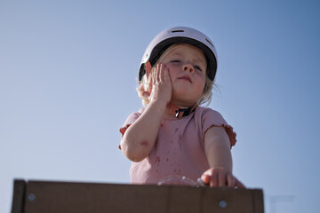 A little girl wearing a helmet drinks water from a public fountain and wipes her face. 