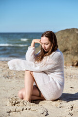 Young woman in white sweater sitting on sandy Baltic Sea beach with calm waves and clear sky