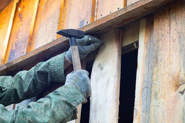 A close-up of a hand wearing a glove, holding a hammer to nail wooden planks on a wall. The scene shows construction work in progress.