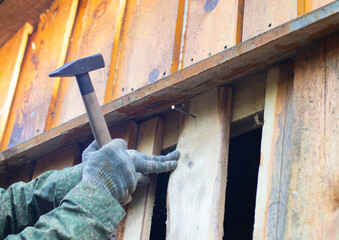 A close-up of a hand wearing a glove, holding a hammer to nail wooden planks on a wall. The scene shows construction work in progress.