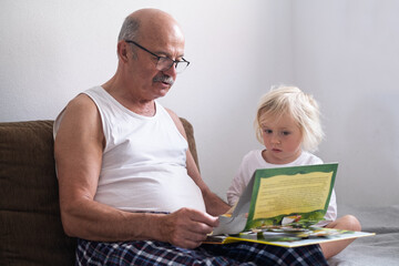 A grandfather is reading a book to his granddaughter while sitting together on a sofa at home. 