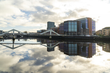 View of modern buildings reflected on the still water under a sky with fluffy white clouds, connected by a bridge, Glasgow, Scotland, United Kingdom.