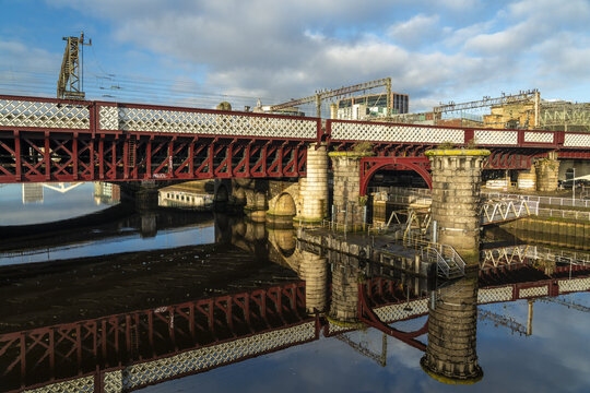 View of intricate red bridge reflected in the dark, rippling water beneath a soft sky, contrasting with the stone pillars, Glasgow, Scotland, United Kingdom.
