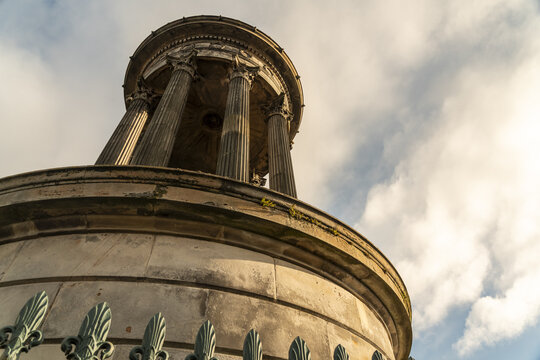 View of Dugald Stewart monument rises against a sky streaked with clouds, framed by a wrought-iron fence, casting long shadows, Calton hill, Scotland, United Kingdom.