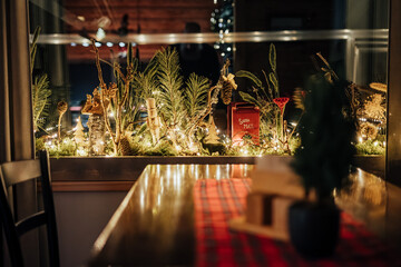 Warm holiday scene with pine branches, fairy lights, and a red Santa Mailbox on a window ledge behind a wooden dining table with copy space