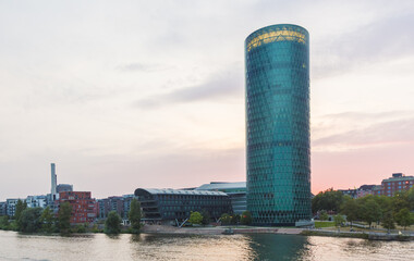 View of a sleek, cylindrical skyscraper reflecting the soft hues of the sunset, contrasting with the calm river, Frankfurt am Main, Hessen, Germany.