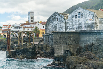 View of weathered stone buildings meet the rugged coastline, a church tower rising amidst colorful buildings, creating a striking contrast, Garachico, Tenerife, Spain.