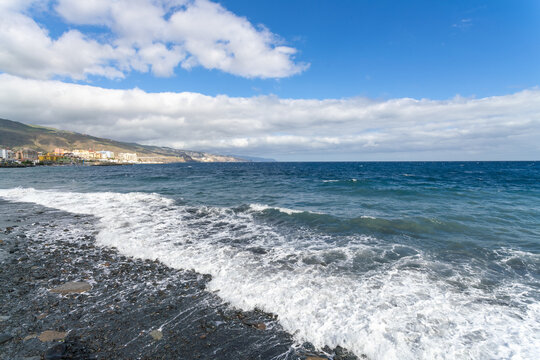 View of foamy waves crash on a dark rocky shore as the turquoise ocean meets a clear sky with fluffy clouds, Tenerife, Canary Islands, Spain.