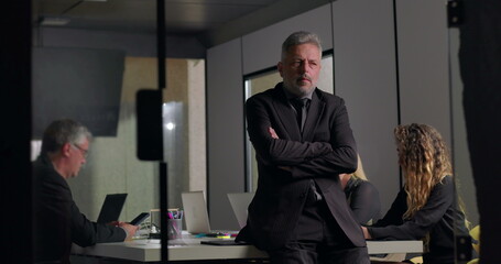 Senior businessman standing with arms crossed and contemplative expression during office meeting while colleagues work on laptops in modern corporate boardroom