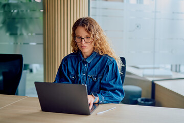 Woman wearing glasses and a denim shirt concentrating on her laptop at a desk in a contemporary office environment