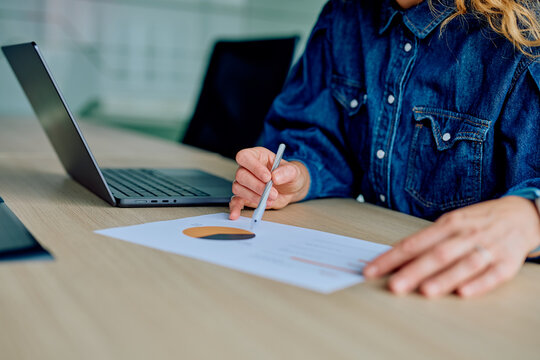 Woman's hand holding a pen, pointing at a chart on a financial report while working on a laptop at a modern office - Powered by Adobe