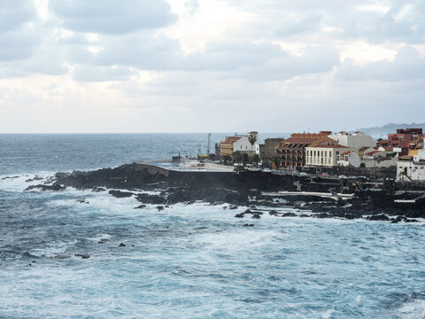 View of the wild, frothy Atlantic Ocean crashing against the dark, jagged volcanic rocks framing the colorful buildings along the coast, Garachico, Tenerife, Spain.