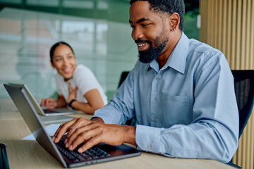 Young businessman typing on laptop, smiling in an office while a female colleague looks on, showing collaboration and teamwork
