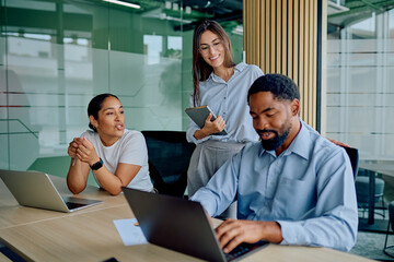 Multi-ethnic business team collaborating at a table with laptops, one woman smiling and observing, showcasing teamwork and productivity