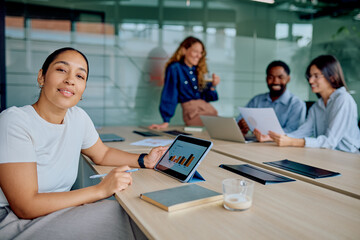 Obraz premium Young businesswoman smiling while using a tablet with charts during a corporate meeting, her colleagues discussing in the background