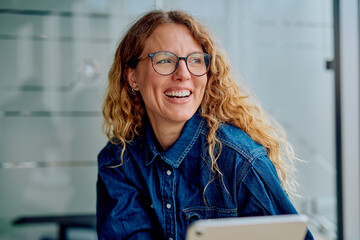 Woman smiling brightly with glasses and curly hair, enjoying a moment while working in a modern...