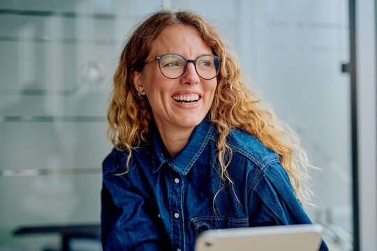 Woman smiling brightly with glasses and curly hair, enjoying a moment while working in a modern office with her tablet - Powered by Adobe