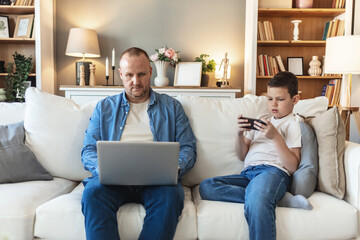 Father using digital tablet and son smart phone while sitting on sofa at home. Father and son bonding at home.