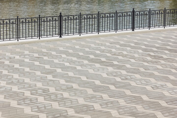 An elegant wrought iron fence borders the shore of a calm river, with a gray sidewalk with a zigzag pattern in the foreground.