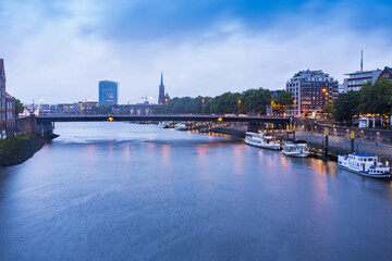 View of a tranquil river reflecting the city lights under a twilight sky, with boats gently floating beneath a bridge in Bremen, Bremen, Germany.