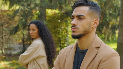 Fototapeta premium Frustrated Hispanic man offended angry separating from upset Arabian woman couple conflict in autumn park date sad boyfriend husband with girlfriend wife outdoors family quarrel ignoring each other