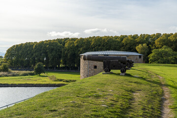 View of a historic cannon standing guard atop a grassy hill near calm waters, with a stone structure and lush trees in the background, Kalmar, Kalmar County, Sweden.