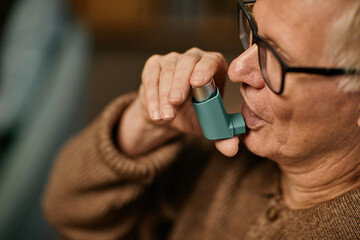 Senior Caucasian man using inhaler while sitting in nursing home, holding device close to mouth,...