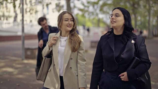 Two confident young businesswomen walk and communicate on a city street, exchanging ideas and discussing plans with smiles. A scene of dynamic urban life and professional interaction.