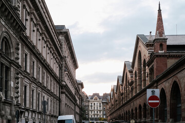 View of aged buildings standing tall in the cityscape, with brick arches and intricate designs contrasting against the overcast sky, Budapest, Hungary.
