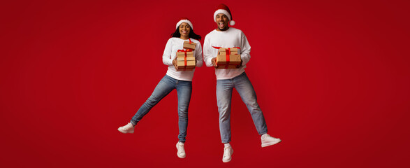 A couple stands together, wearing Santa hats and smiling broadly while holding wrapped gifts. Their...