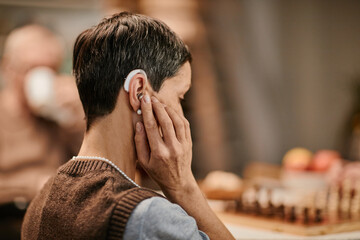 Senior Caucasian woman adjusting hearing aid while sitting in nursing home, short hair visible,...