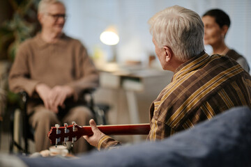 Senior Caucasian man playing acoustic guitar for senior Caucasian man with disability in wheelchair, middle aged Caucasian woman sitting together in nursing home common area