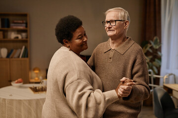 Senior Black woman and senior Caucasian man dancing together in nursing home, holding hands and smiling, enjoying companionship and connection in comfortable indoor setting