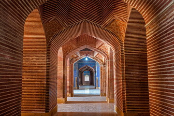 View of a long corridor of red brick arches adorned with geometric patterns, leading to a distant light, in Thatta, Sindh, Pakistan.