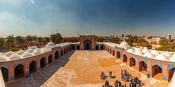 View of an expansive courtyard adorned with arched walkways and domes, where a group of people gathers under the bright sky, Thatta, Sindh, Pakistan.