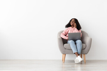 A young woman sits in a cozy chair, smiling while using her laptop and talking on a phone. The room is bright and minimalistic, creating a relaxed atmosphere.
