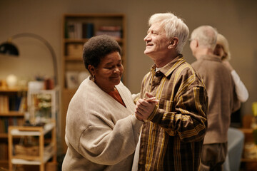 Senior Black woman and senior Caucasian man dancing together in nursing home, smiling and holding hands, another senior couple interacting in background, warm indoor setting