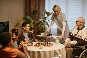 Group of senior Caucasian man and woman and Black woman sitting around table playing chess, while young adult Caucasian female caregiver standing nearby assisting in nursing home setting