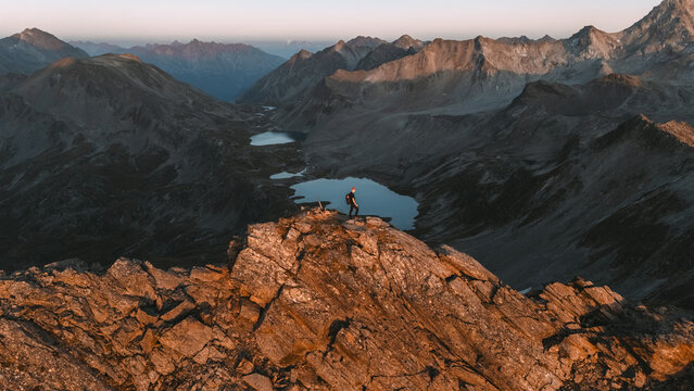 View of a lone figure standing atop jagged, sun-kissed peaks overlooking tranquil, reflective lakes nestled amongst the vast, rugged mountains, Klosters-Serneus, Grisons, Switzerland.