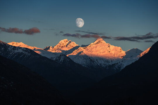 View of snow-capped peaks kissed by the warm glow of the setting sun, contrasting against deep shadows under a bright moon, Zermatt, Valais, Switzerland.