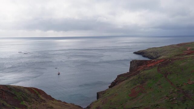 Aerial view of Ponta de Sao Lourenco on Madeira, steep cliffs meet calm Atlantic, two sailboats anchor near circular fish pens, overcast light, narrow trail visible.