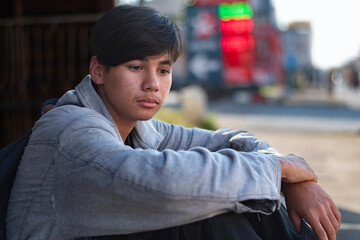 A somber middle schooler, dressed in grey and black, waits alone at the gas station. The black backpack and downcast face capture a moment of teenage melancholy, transit, and quiet introspection.