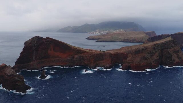 Aerial view shows ochre red cliffs and sea stacks at Ponta de Sao Lourenco, Madeira, with waves on a rocky shore, distant Canical, white towers, and overcast light.
