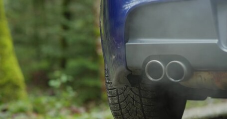 Generic car twin exhaust pipe tips emitting gray smoke or condensation while revving the engine. Low angle, close up shot, blurry nature background, dynamic track and pan real time video, no people