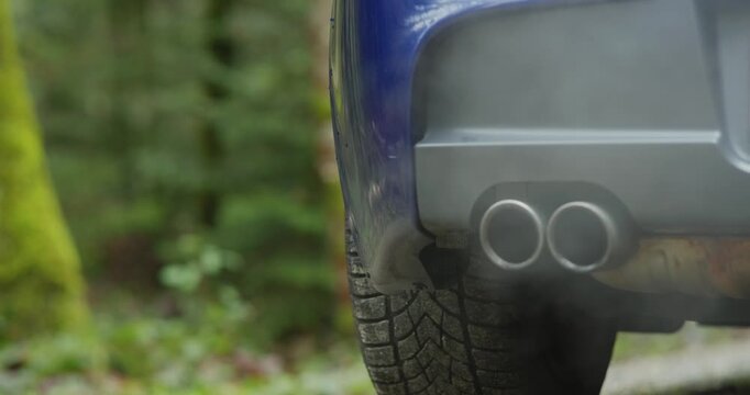 Generic car twin exhaust pipe tips emitting gray smoke or condensation while revving the engine. Low angle, close up shot, blurry nature background, dynamic track and pan real time video, no people