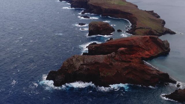 Aerial view shows Ponta de Sao Lourenco, Madeira, Portugal. Indigo Atlantic waves churn around sea stacks and coves as the camera sweeps slowly in overcast light.