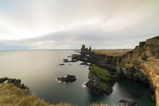 View of dramatic cliffs plunge into the still, emerald sea under a soft, diffused sky, where rugged textures meet serene waters, Hellnar, Snaefellsbaer, Iceland.