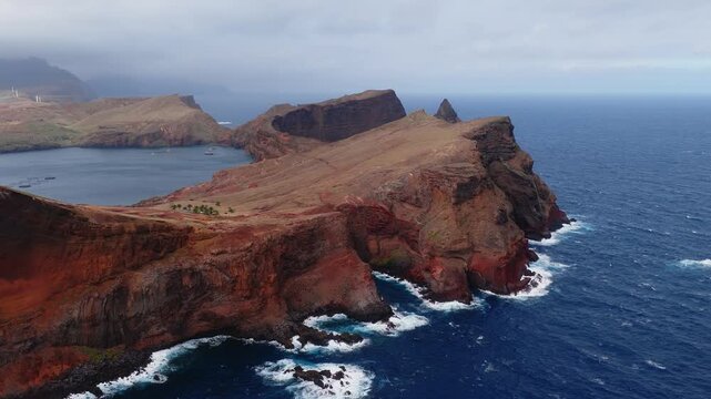 Aerial view of Ponta de Sao Lourenco, Madeira, Portugal, as a drone glides past red black cliffs, sea stacks, cliff caves, fish pens, a bay, and distant wind turbines in overcast light.