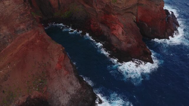 Aerial camera glides along Madeira cliffs as waves strike basalt coves, layered erosion and sparse green patches visible in low light with rich color and scale.