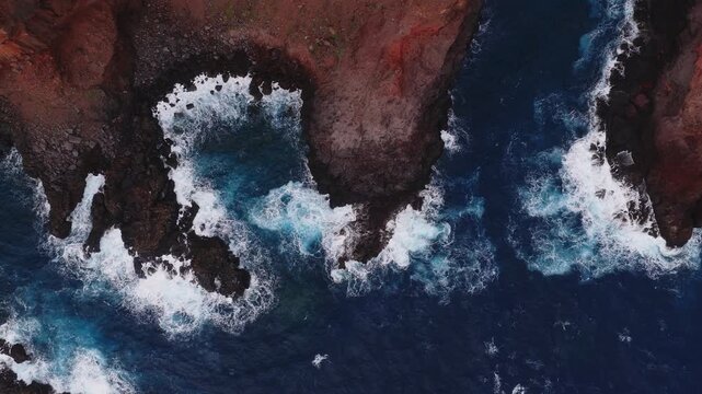 Aerial view shows rugged red cliffs on Madeira, Portugal, as waves crash into coves and sea caves. Overhead pans and top down passes reveal warm light and cool blues.